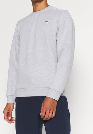 Man draagt een lichtgrijze trui met lange mouwen en een klein groen krokodillenlogo op de borst, gecombineerd met donkerblauwe broek, staand tegen een witte achtergrond.