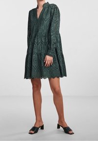 Dunkelgrünes Kleid aus Spitze mit V-Ausschnitt und langen Ärmeln. Verfügt über einen ausgestellten Saum mit gewelltem Rand und kunstvollen Ausschnittmustern.