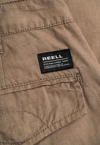 Beiger Stoff mit strukturiertem Karomuster, Taschennaht und schwarzem Label mit der Aufschrift "REELL Established 1997 Engineered by United People."