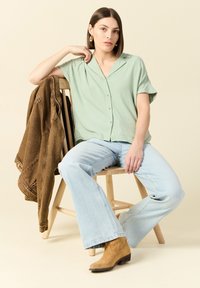 Lichtgroen overhemd met korte mouwen, vervaagde blauwe flared jeans, bruine denim jack over een houten stoel, tan enkellaarzen.