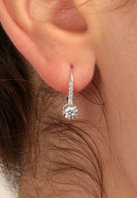 Nahaufnahme eines Ohrläppchens mit einem silbernen Tropfenohrring, der eine Reihe kleiner Diamanten und einen größeren runden Diamanten am Ende hat.