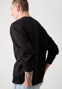 Sweatshirt negru cu guler rotund și mâneci lungi, dotat cu manșete ribbate. Textură netedă, croi casual și design pe spate prezentat.