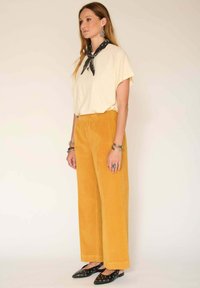 Haut court jaune clair à manches courtes, pantalon large en velours côtelé moutarde, chaussures noires à talons pointus avec des œillets, écharpe noire nouée autour du cou.