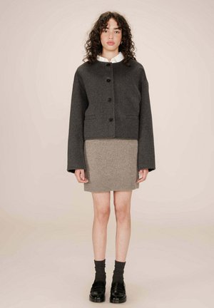 Veste en laine grise, courte, avec un col rond, cinq boutons et deux poches au niveau des hanches, associée à une jupe tricotée côtelée marron clair.