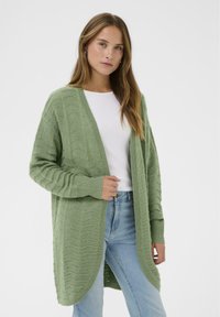 Groene gebreide cardigan met een gestructureerd golfpatroon, een open voorkant, lange mouwen en geribbelde manchetten. Draagt over een wit shirt en blauwe spijkerbroek.