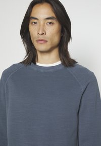 Blauer, gerippter Sweatshirt mit rundem Ausschnitt und Raglanärmeln. Die Textur ist leicht strukturiert und weich. Ein schlichtes weißes Unterhemd ist sichtbar.