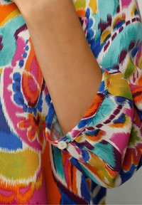 Bras partiellement couvert par une chemise à manches longues colorée et à motifs, avec un poignet boutonné dans des couleurs vives bleu, orange, rose et vert.