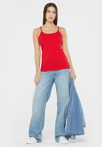 Red sleeveless tank top, fitted design, paired with light blue wide-leg jeans, holding a denim jacket, set against a plain white background.