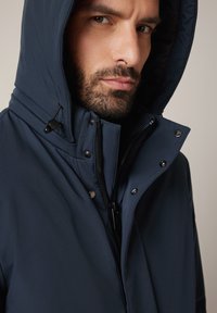 Mężczyzna z brodą, ubrany w granatową kurtkę z kapturem, z zamkiem błyskawicznym i detalami na zatrzask, lekko spogląda w bok.