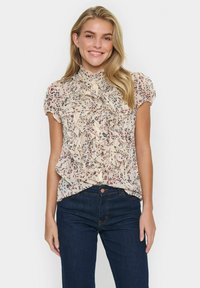 Light beige blouse with a high ruffled neckline, short sleeves, and floral print in shades of red and blue. Worn with dark jeans.