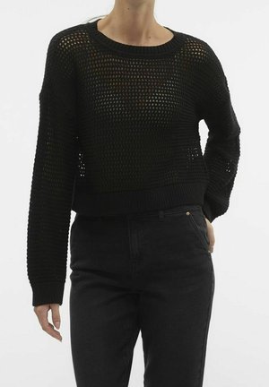 Femme portant un pull en maille noir ample à manches longues et un pantalon noir taille haute, debout avec une main dans la poche.