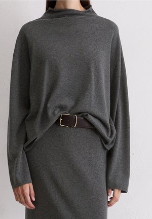 Mujer que lleva un top de manga larga suelto de color gris oscuro metido en una falda a juego con un cinturón marrón oscuro con hebilla dorada.