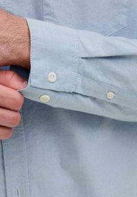 Camicia button-up azzurra chiara, realizzata in tessuto morbido. Presenta un polsino con un solo bottone e un orlo curvo e ordinato. Anche i punti di cucitura sono visibili.