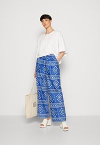 Pantalons larges à motifs bleus avec des designs géométriques blancs ; portés avec un haut blanc ample et des chaussures à talons blocs blancs. Tient un sac fourre-tout beige.