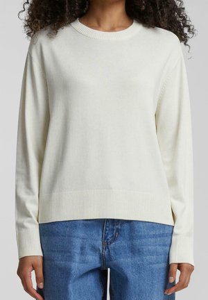 Femme portant un pull crème uni à col rond avec des poignets et un bas côtelés, associé à un jean bleu à lavage moyen.