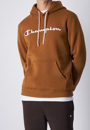 Homme portant un sweat à capuche Champion marron avec cordons blancs et logo, associé à un pantalon foncé avec un petit logo Champion.