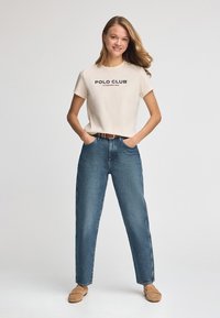 Krämvit T-shirt med korta ärmar och texten "POLO CLUB"; matchas med lösa blå jeans och beigefärgade slip-in skor. Enkel, avslappnad design.