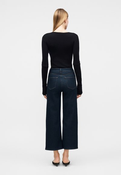 Γυναίκα με μακριά ξανθά μαλλιά στέκεται γυρισμένη προς τα πίσω, φοράει μαύρο τοπ με μακριά μανίκια, φαρδιά σκουρόχρωμα μπλε τζιν και μαύρες γόβες.