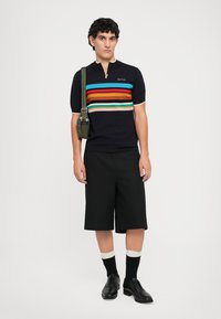 Lyhythihainen tummansininen polo, jossa on monivärisiä vaakaraitoja, mustat shortsit, mustat kengät ja vihreä olkalaukku. Pehmeä neulekangas.