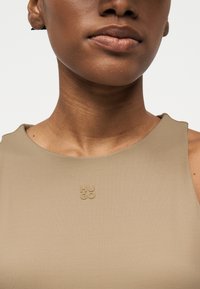 Κοντινό πλάνο του λαιμού και του κάτω μέρους του προσώπου ενός ατόμου που φοράει μπεζ αμάνικη μπλούζα με μικρό ανάγλυφο λογότυπο «HUGO» στο στήθος.