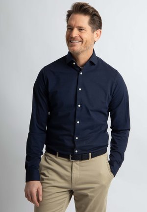 Marineblauw overhemd met lange mouwen in een aansluitende pasvorm, knoopsluiting aan de voorkant en witte knopen. Gecombineerd met beige broek en een zwarte riem.