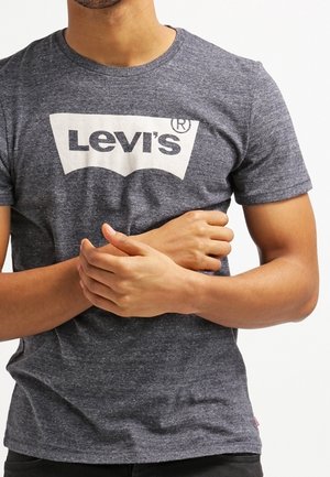 Mężczyzna w szarej koszulce Levi's ze skrzyżowanymi rękami, widoczny od szyi do bioder, na jednolitym białym tle.