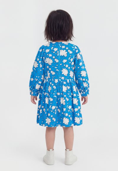 Dieťa stojí chrbtom, oblečené v modrých šatách s bielymi vzormi oblakov, hviezd a mesiaca a bielych členkových topánkach.