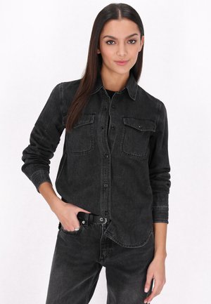 Zwart denimshirt met twee borstzakken, knoopsluiting, lange mouwen en een licht vervaagde textuur. Gecombineerd met bijpassende zwarte jeans.
