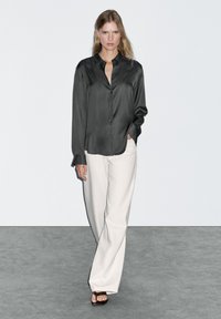 Camicia nera di satin con maniche lunghe, colletto e bottoni, abbinata a pantaloni bianchi a gamba larga e sandali marroni, su uno sfondo semplice.