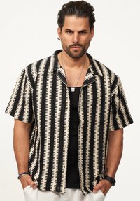 Kurzarmhemd mit einem geometrischen Muster aus schwarz-beigen Streifen, Knopfleiste und leichtem Stoff. Auffällige strukturierte Details sind enthalten.