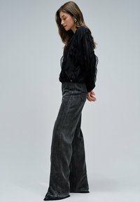 Blusa trasparente nera con volant abbinata a jeans grigi a gamba larga e vita alta. Tessuto texturizzato con dettaglio di spacco sull'orlo della gamba.