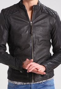 Giacca in pelle nera con design trapuntato sulle spalle, collo alto e cerniere frontali e sui polsini. Vestibilità slim con dettagli sottili.