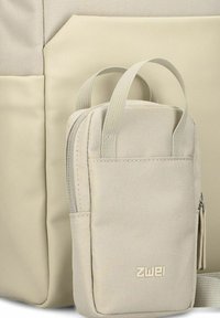 Zaino di tessuto beige con una piccola tasca staccabile. Il materiale è texturizzato, con un pannello frontale liscio e un branding sottile sulla tasca.