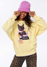 Žena v žluté mikině s černou kočkou a nápisem "Káva a žádné plány", růžové čepici a kalhotách s leopardím vzorem, usmívající se se zavřenýma očima.