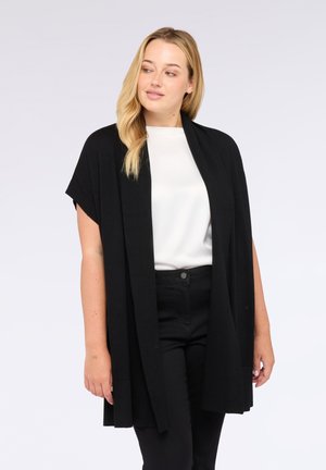 Femme aux cheveux blonds portant un haut blanc, un cardigan sans manches noir et un pantalon noir, regardant légèrement vers sa gauche contre un fond uni.