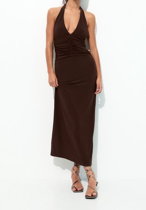 Kobieta w bezrękawowej, ciemnobrązowej sukience midi z marszczeniami i głębokim dekoltem w kształcie litery V, nosząca srebrne sandały na wysokim obcasie na cienkich paseczkach na białym tle.