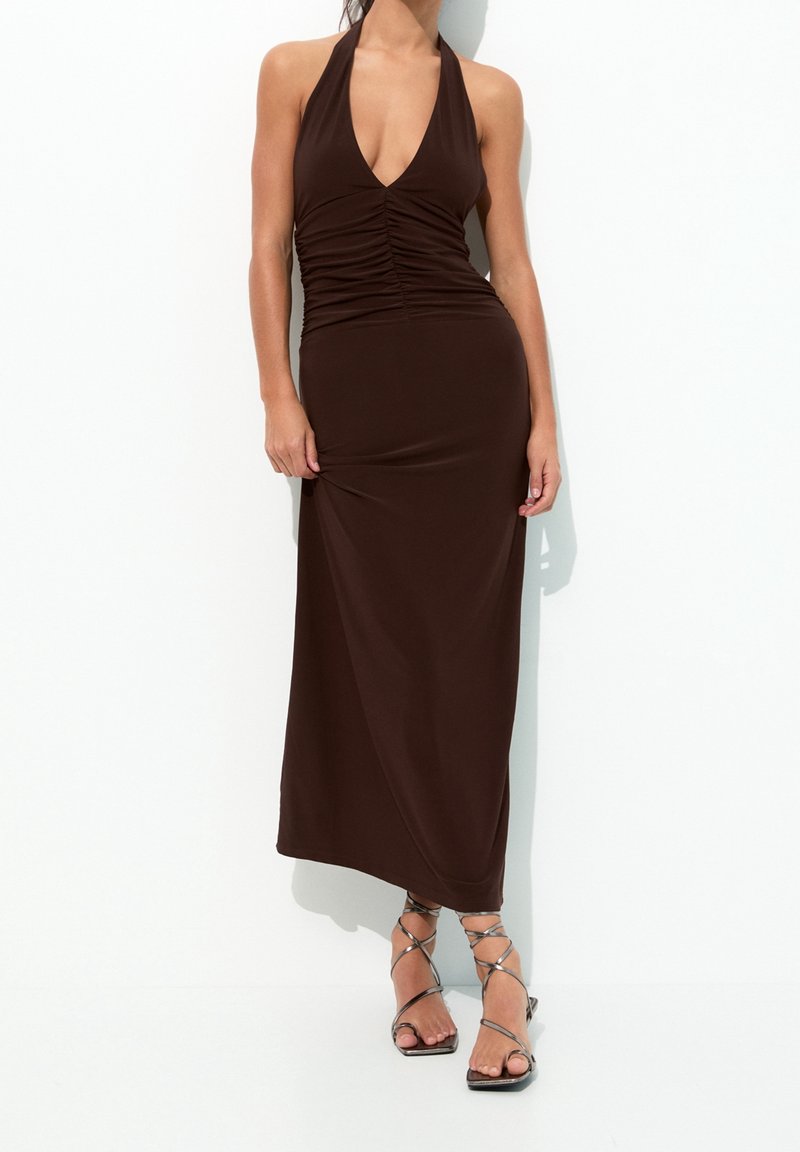 Femme portant une robe midi marron foncé froncée, sans manches et avec un décolleté en V profond, ainsi que des sandales à talons hauts argentées à brides, sur fond blanc.