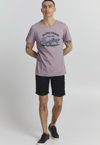 Lavendelfarget crew neck t-skjorte med svart og blå "Blend Denim Surfing" grafikk, kombinert med svarte rullede shorts og svarte sneakers.
