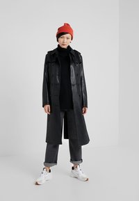 Zwarte leren trenchcoat met een dubbelzijdig ontwerp, grote kraag en zijzakken. Gecombineerd met donkere jeans, witte sneakers en een rode beanie.