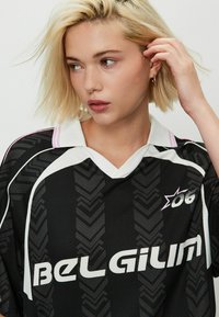 Ung blond kvinna som bär en svart sporttröja med texten "Belgien", tittar åt sidan och rör vid sitt hår mot en enfärgad bakgrund.