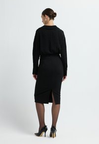 Schwarze Bluse mit strukturiertem Muster, kombiniert mit einem taillierten schwarzen Rock mit einem Schlitz hinten. Das Model trägt schwarze High Heels und transparente Strumpfhosen.