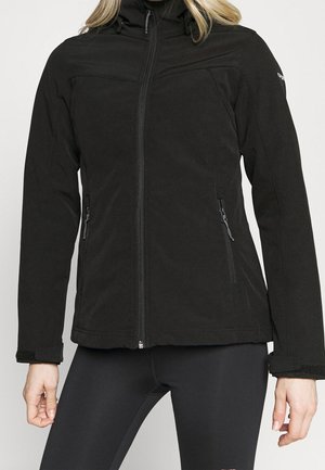 Frau trägt eine taillierte schwarze Jacke mit Reißverschluss und Reißverschlusstaschen an den Seiten sowie langen Ärmeln, kombiniert mit schwarzen Leggings.
