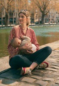 Gestreepte rood-witte blouse, blauwe spijkerbroek, zittend op kasseien bij het water. Houdt een baby vast in een beige trui, draagt meerkleurige sneakers.