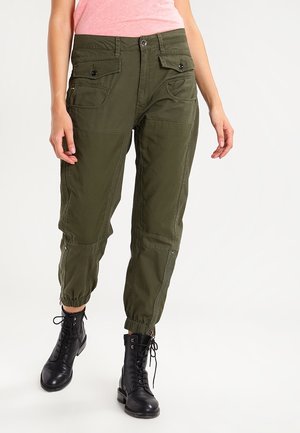 Kvinde iført olivengrønne cargobukser med lommer med knapper og sorte snørestøvler, stående mod en ensfarvet baggrund.