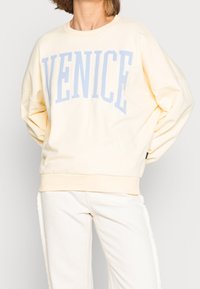 Felpa giallo chiaro con la scritta “VENICE” in blu sul petto, caratterizzata da un colletto rotondo e maniche a vestibilità ampia, abbinata a pantaloni bianchi.