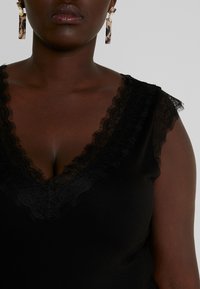 Top nero con scollatura a V profonda e bordi in pizzo, caratterizzato da eleganti edge arricciati. Accentuato da orecchini a cerchio e una texture di tessuto liscia.