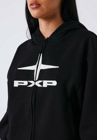 Femme portant un sweat à capuche noir zippé avec un logo en forme d'étoile blanche et le texte "PXP" sur la poitrine, vue des épaules aux lèvres.