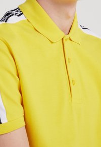 Polo jaune avec un tissu texturé, manches courtes et col classique ; présente des accents noirs et blancs sur les épaules.