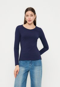 Γυναίκα με μακριά καστανά μαλλιά που φορά χρυσαφένια σκουλαρίκια κρίκους, μπλε μακρυμάνικο μπλουζάκι και ανοιχτό μπλε τζιν, στέκεται μπροστά σε ένα λευκό φόντο.