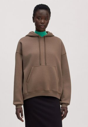 Young woman wearing a loose brown hoodie over a green turtleneck and a black skirt, standing against a plain background.